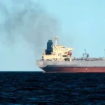 Большой серый танкер с дымом и военный корабль на море