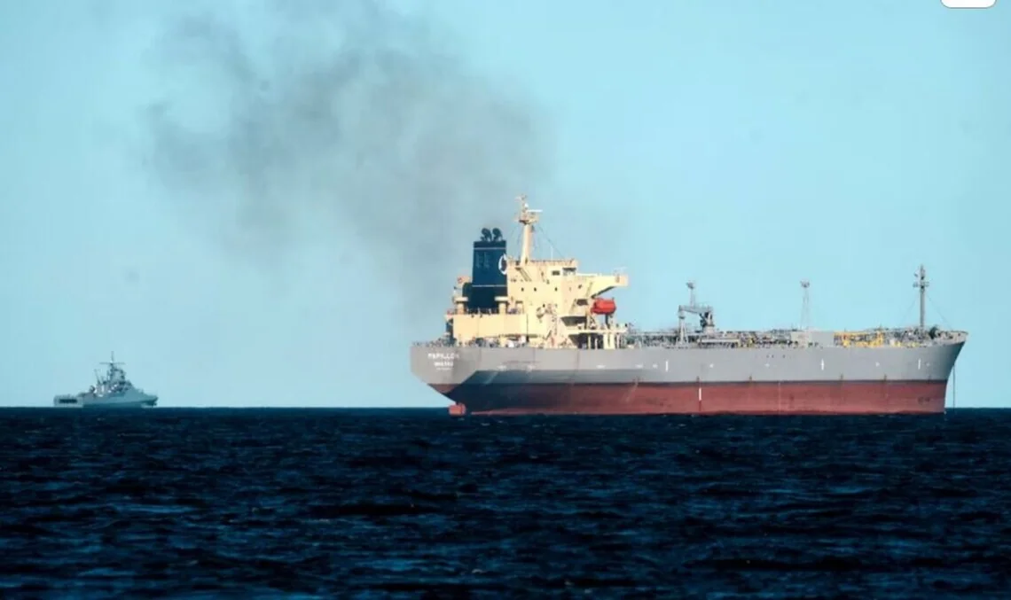 Большой серый танкер с дымом и военный корабль на море