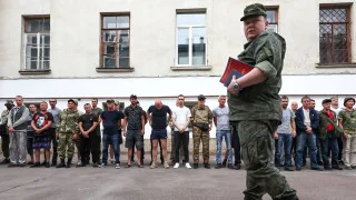 Офіцер у формі з червоною папкою йде повз шеренгу чоловіків біля будівлі